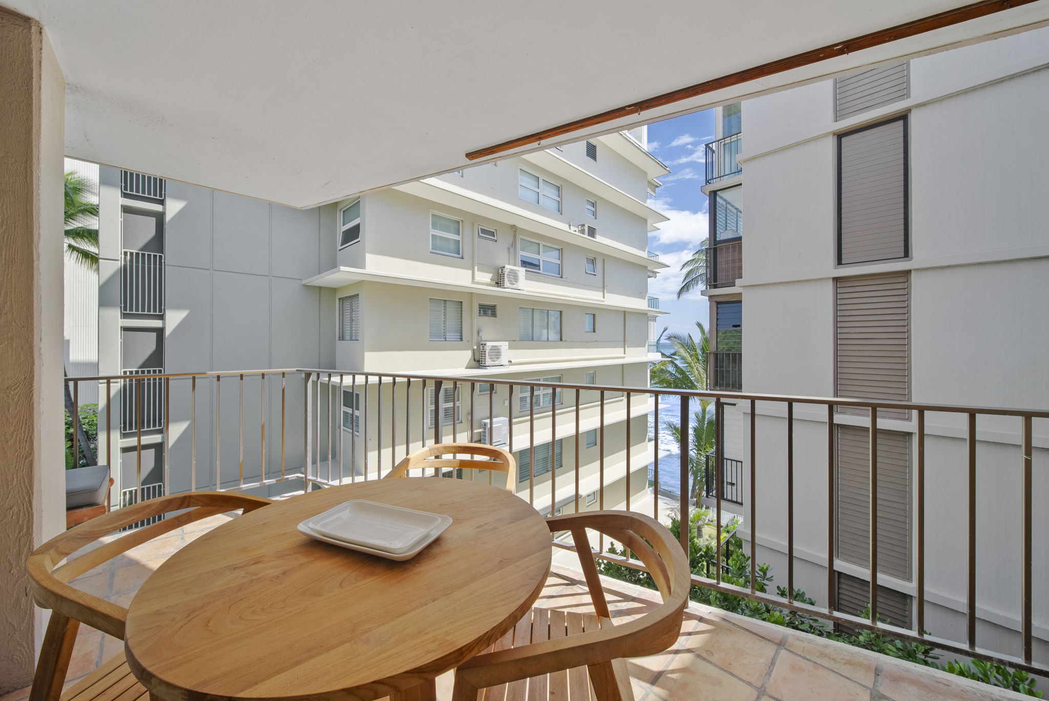Sunny lānai with a wood dining table and a glimpse of the Pacific between neighboring buildings